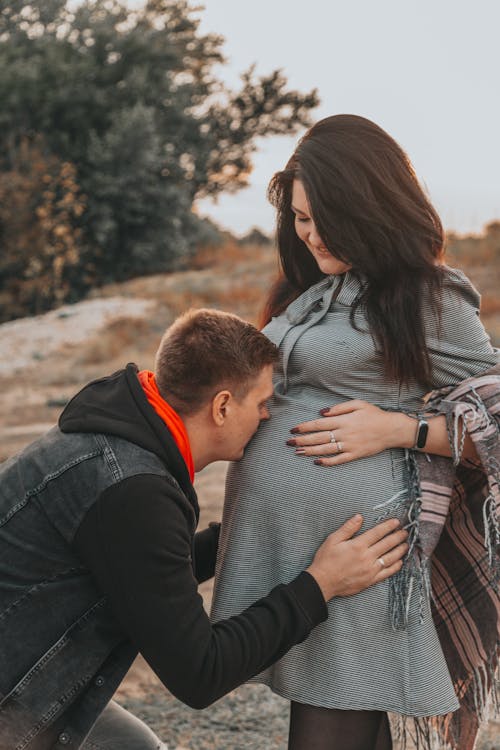 Side view of young man in casual outfit hugging and kissing pregnant wife with long hair in belly