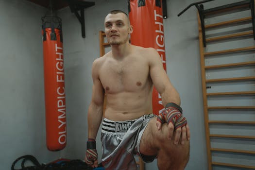 A shirtless muscular man in gray shorts with hand wraps in a gym setting.