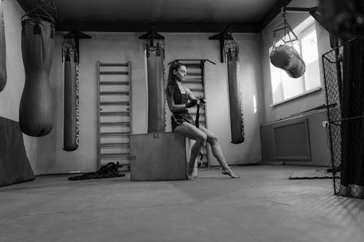 A woman prepares for a workout in a monochrome gym setting, surrounded by punching bags and gym equipment.