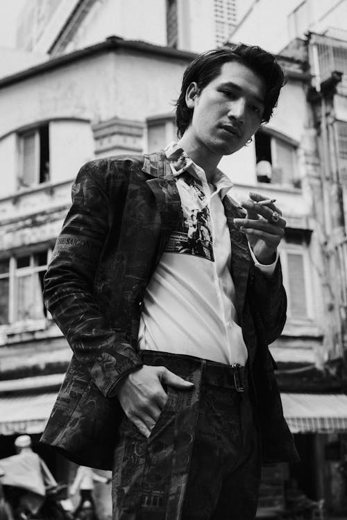 Low angle of black and white young confident ethnic guy standing on street near aged buildings with hand in pocket and smoking cigarette