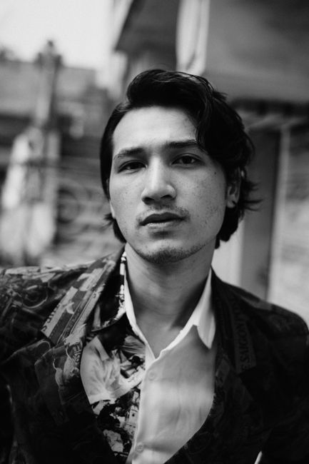 Black and white of calm young ethnic guy with dark wavy hair in casual suit looking at camera while resting on city street