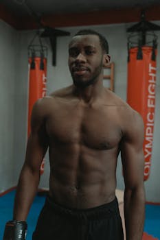 Muscular man standing shirtless in a gym with punching bags in the background.
