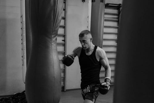 Black and white photo of a muscular male boxer training with punching bags indoors.