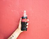 A tattooed hand holding a Coca-Cola bottle against a vibrant red wall in Puerto Rico.