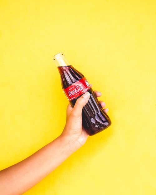 Free Close-up of a hand holding a Coca-Cola bottle with a vibrant yellow backdrop, creating a bright and engaging image. Stock Photo