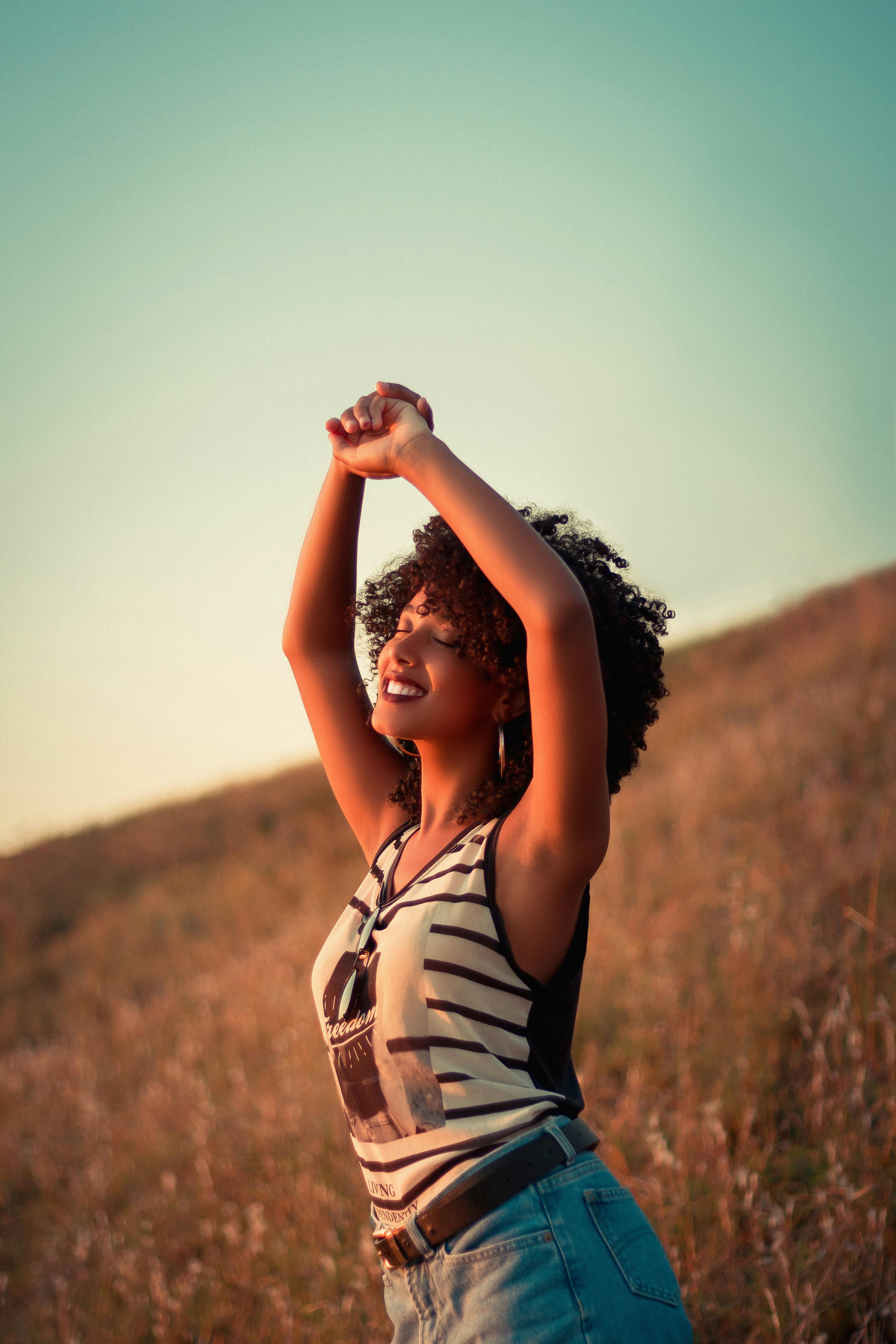 一位面带微笑的年轻非裔美国女性，穿着牛仔裤和条纹背心，闭着眼睛举起双手，享受大自然