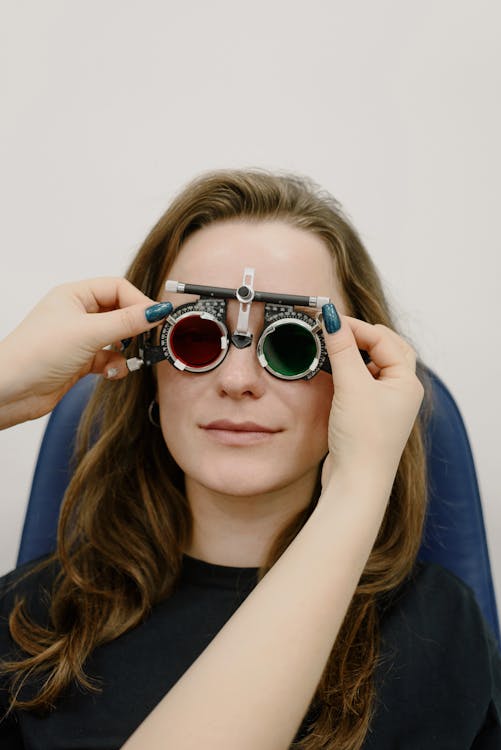 Unrecognizable oculist correcting phoropter with tinted lenses on eyes of female patient while doing vision check up against white wall