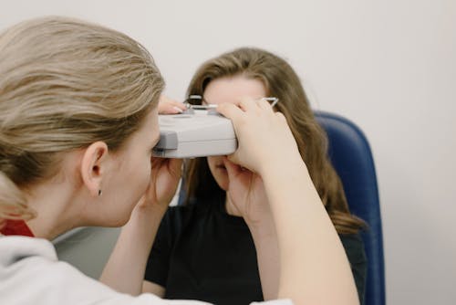 Anonymous female patient looking at optical microscope and sitting near white wall near oculist while making vision diagnostic during appointment in clinic