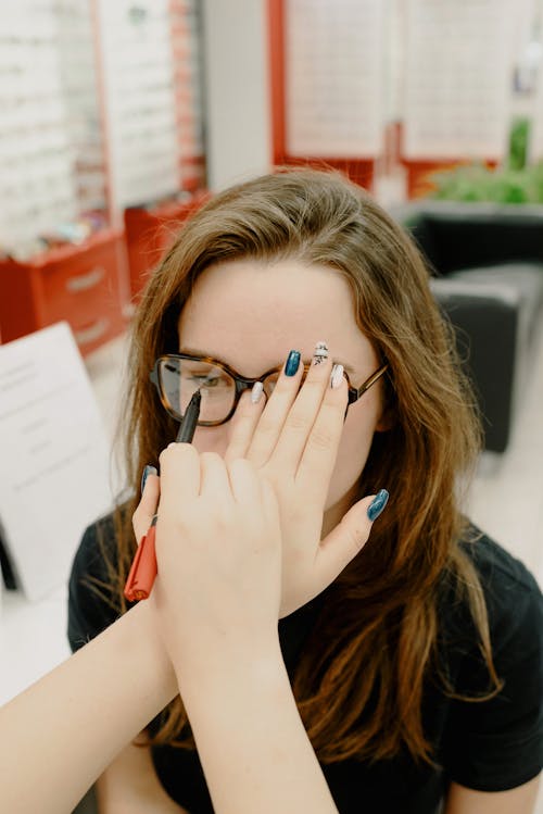 From above of unrecognizable ophthalmologist testing vision of female patient while standing with pen and covering eye in clinic on blurred background