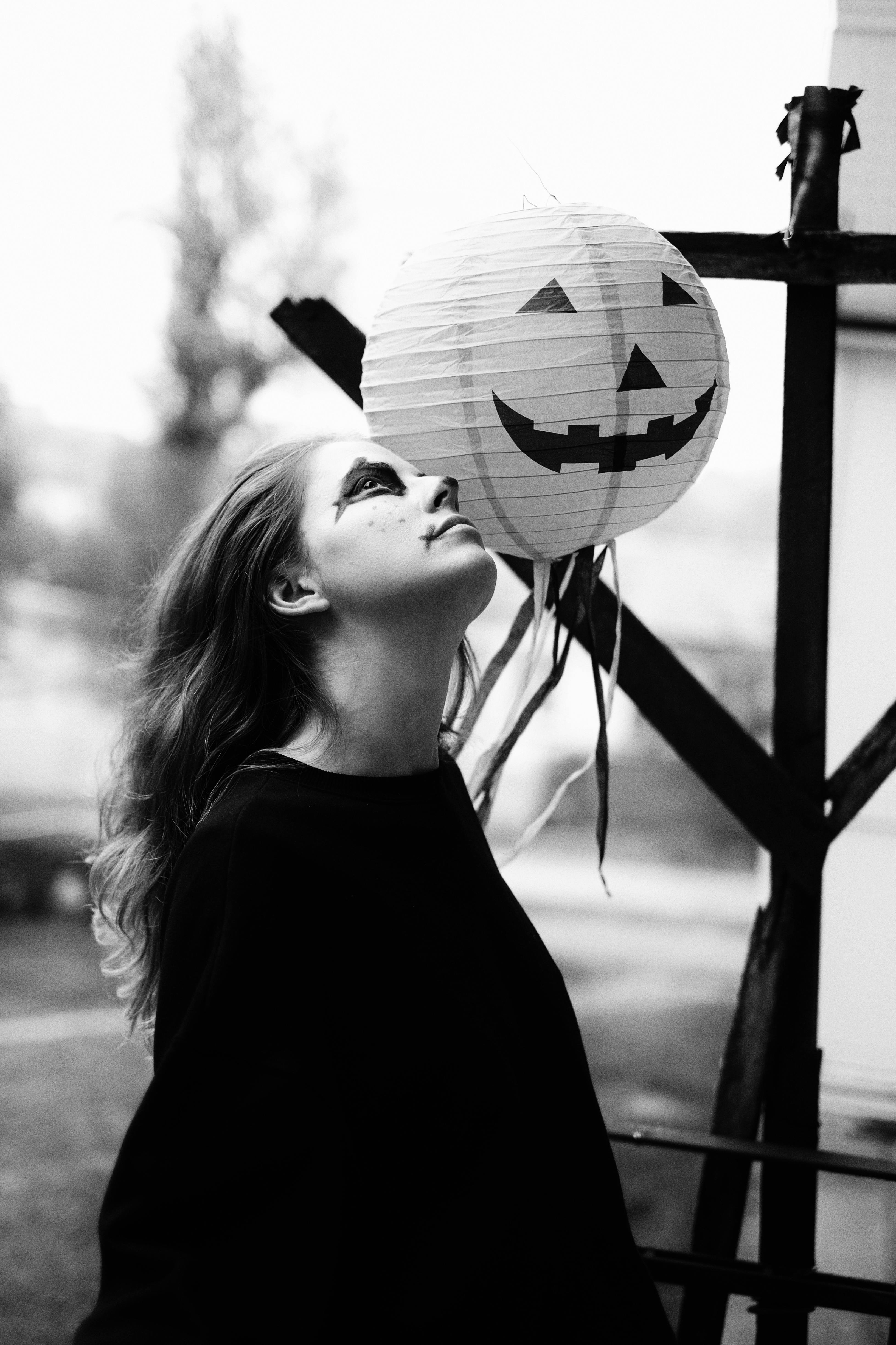 A young woman in creative makeup looks up at a jack o' lantern paper lantern outdoors.
