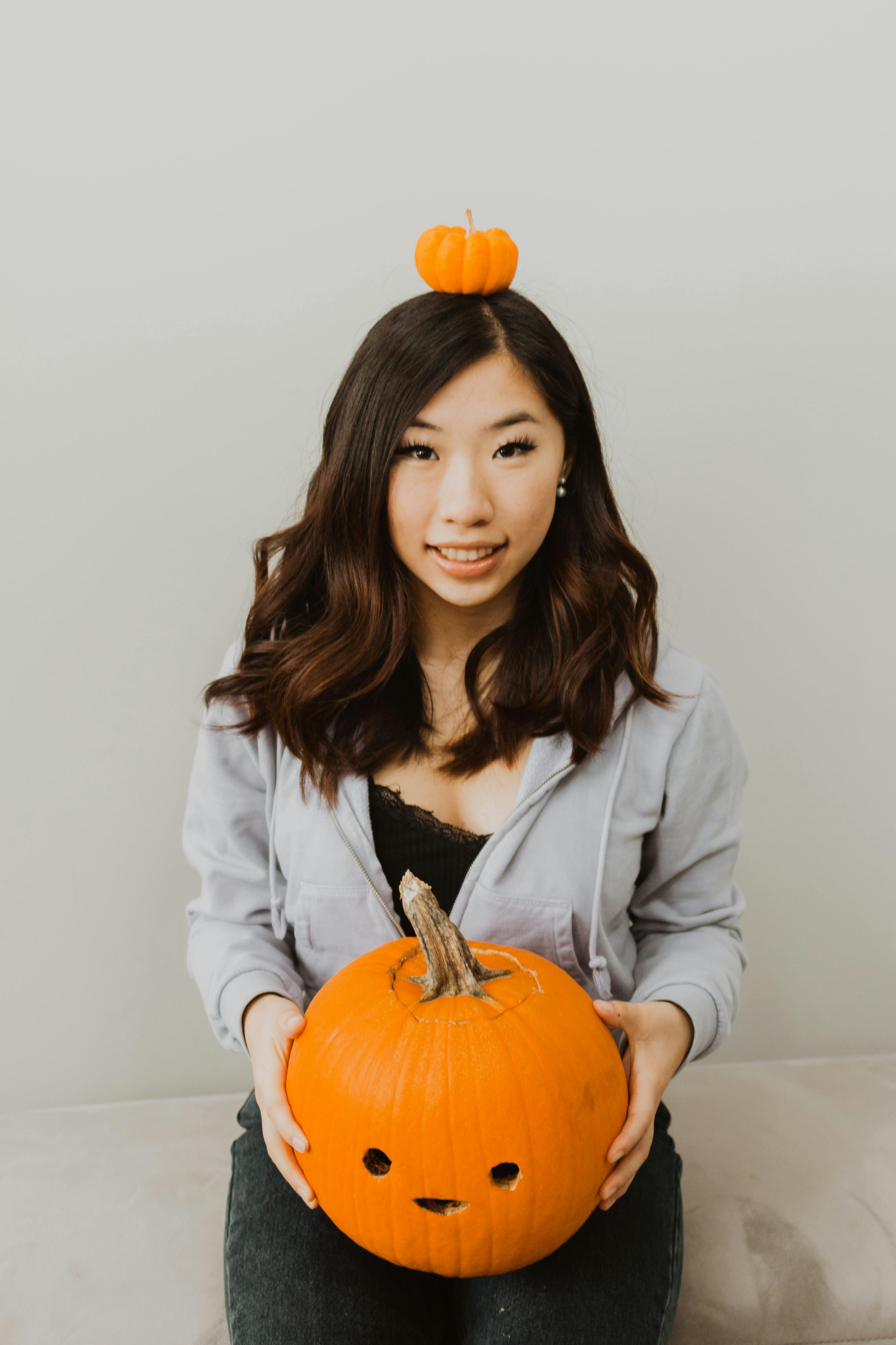 Cheerful Asian female with dark hair looking at camera while sitting on coach with decorative pumpkin on head and carved jack o lantern