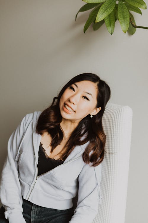 Free Delighted Asian lady with dark hair with toothy smile looking at camera while sitting on chair near wall with head bowed Stock Photo