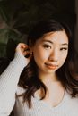 Attractive Asian female with dark hair wearing earring looking at camera while tucking hair behind ear and sitting near green plant