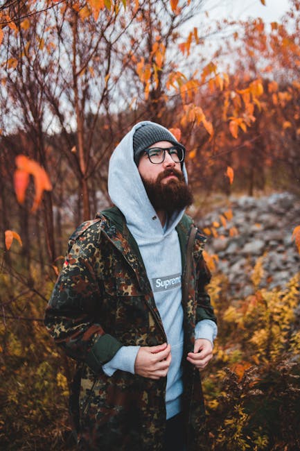 Bearded hipster male in eyeglasses standing among trees with autumn foliage and looking up