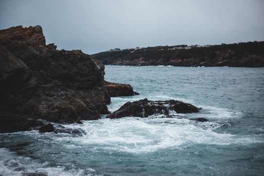 Spectacular scenery of foamy waves of powerful sea crashing near rough rocky cliff against misty gray sky