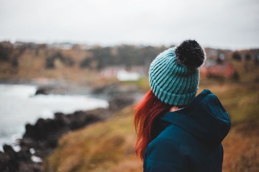 Side view of unrecognizable young female hiker with long red hair in warm outerwear and hat admiring stormy ocean with rocky coast from grassy hill against cloudy sky