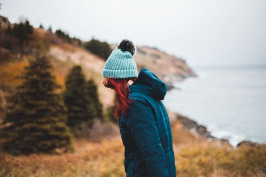 Side view of female in warm clothes standing on lawn with yellow grass and coniferous trees on blurred background of river