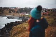 Back view of female in outerwear admiring cottages of countryside on coast covered with grass