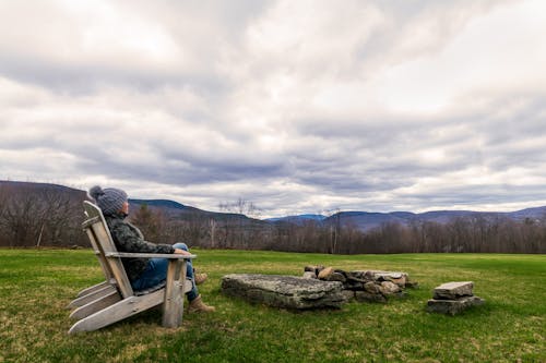 Side view of relaxed young female traveler in warm outerwear and hat sitting on wooden chair on green grassy meadow and admiring picturesque mountainous landscape against cloudy sky