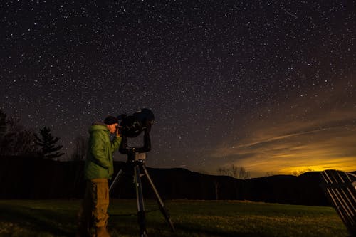 Side view of unrecognizable male traveler in warm clothes and hat observing starry night sky with telescope standing on grassy meadow in hilly terrain