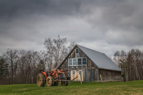 Free Exterior of weathered wooden house and dirty tractor on grassy field near forest with leafless trees against dramatic overcast sky in farmland Stock Photo