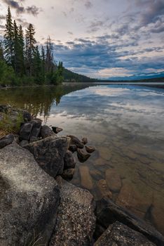 Calm lake with rocky shoreline in Jasper National Park, showcasing tranquil reflections of trees and cloudy sky.
