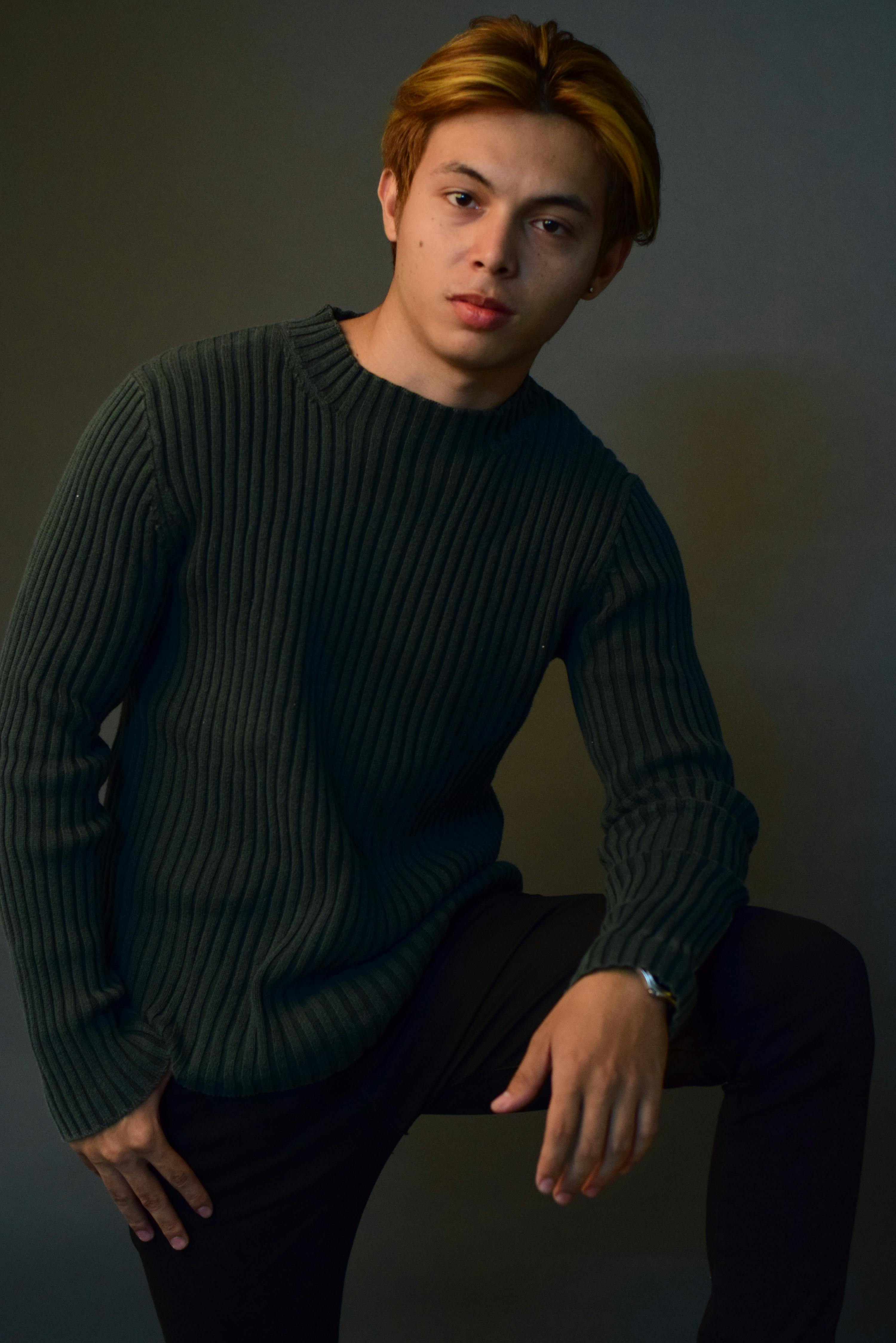 A young man in a knitted sweater poses with a serious expression in a studio setting.