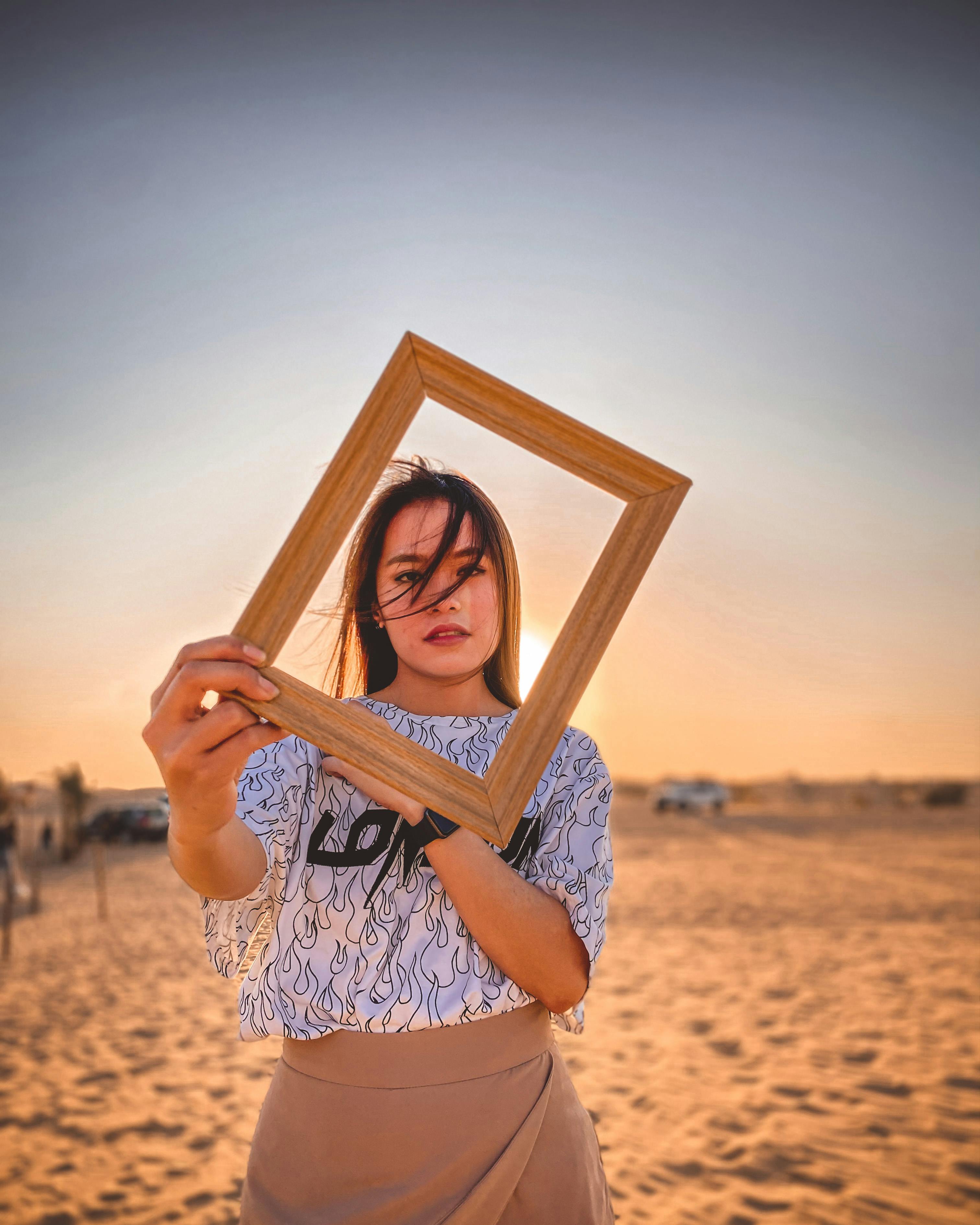 Young ethnic female in casual clothes holding wooden frame in front of face and looking at camera while relaxing on sandy beach against cloudless sunset sky