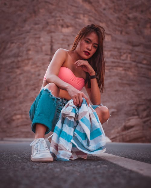 Confident Asian female with long hair in stylish outfit squatting down on asphalt road against rocky slope of mountain