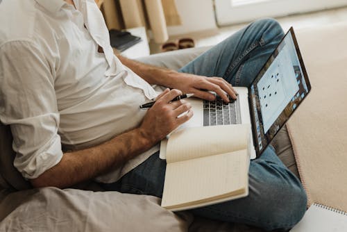 Orang Dengan Kemeja Putih Dan Jeans Denim Biru Yang Duduk Di Tempat Tidur Menggunakan Macbook Pro