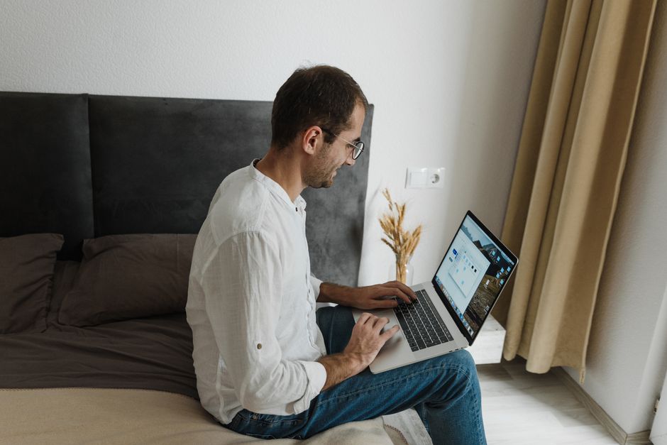 How Hybrid Mattresses Alleviate Back Pain for Chronic Sufferers A man in white shirt working on a laptop while sitting on his bed, depicting home office lifestyle.