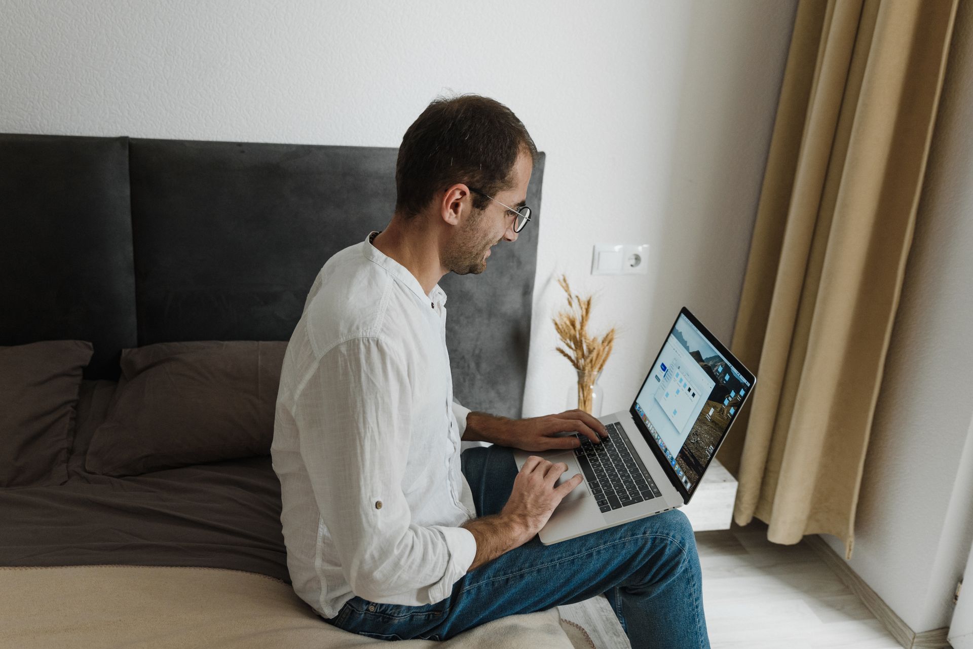 A man in white shirt working on a laptop while sitting on his bed, depicting home office lifestyle.