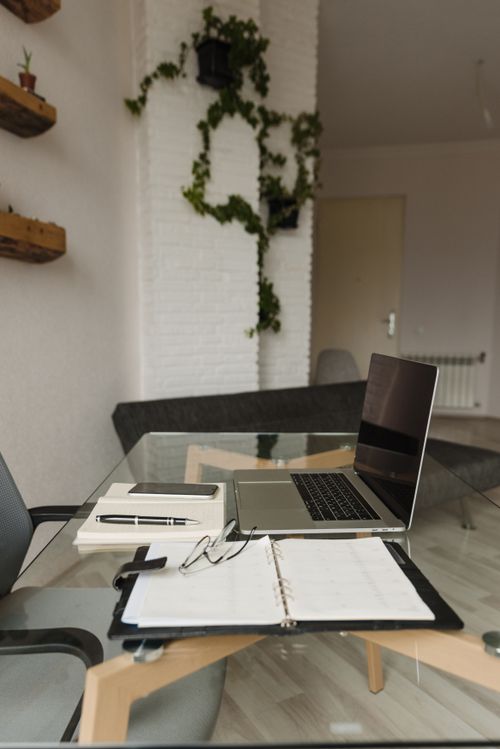 Free Cozy home workspace featuring a laptop, planner, and glass table with plants. Stock Photo