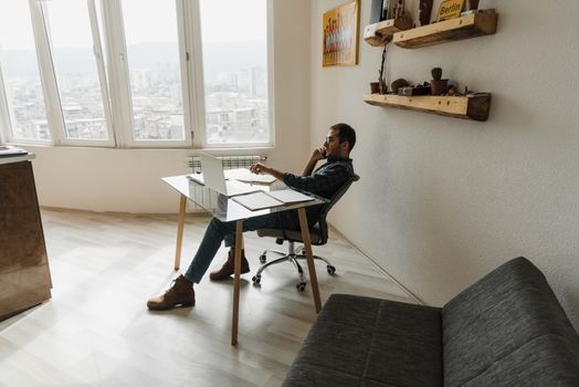 Man on phone working remotely from home office with laptop and city view.