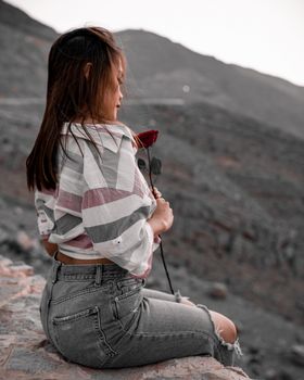 一个女人坐在岩石山上，手里拿着一朵红玫瑰，背景宁静