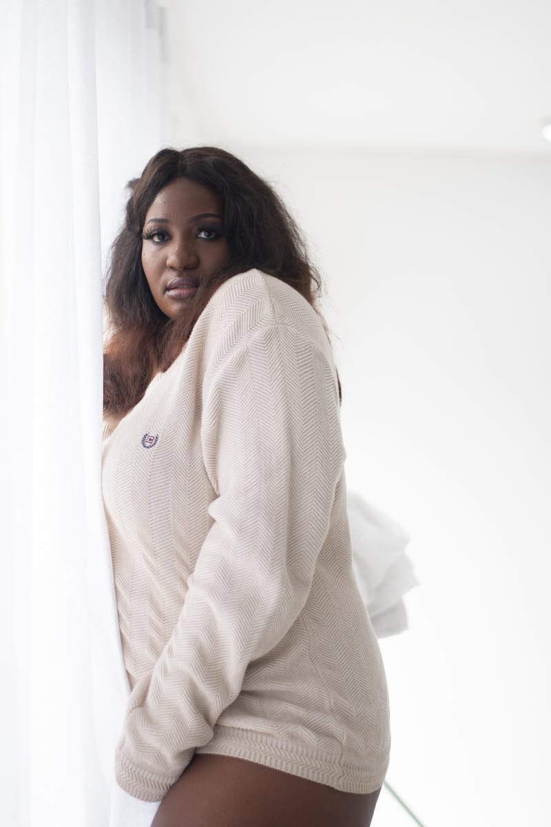 Portrait of a stylish woman in a white sweater standing by a bright window indoors.