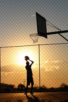 Silhouetted athlete playing basketball on an outdoor court with a vibrant sunset backdrop.