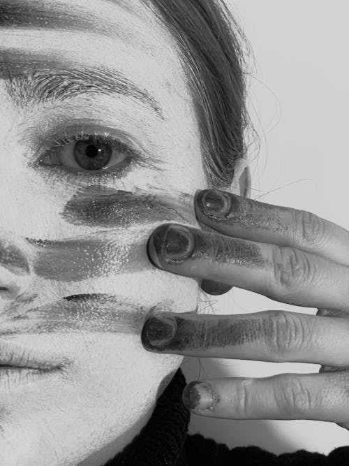 Crop black and white melancholic female smearing dark paint on face with artistic makeup and looking at camera