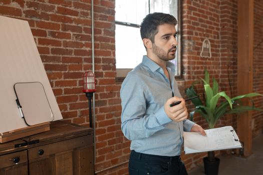 A young man stands in a loft with brick walls teaching or presenting with a clipboard and pen.