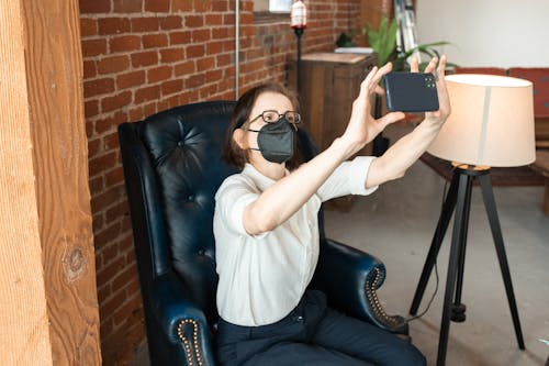 A Person in a Face Mask Taking a Selfie while Sitting in an Armchair