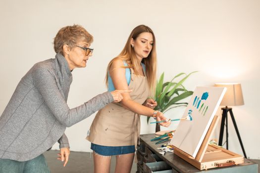 Two women collaborate in an art class, painting on canvas indoors.