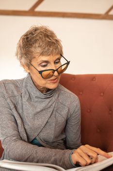 一位戴着眼镜的女士坐在红色沙发上，穿着灰色服装，在室内读书