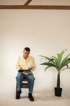 一位穿着黄衬衫的成年男子坐在凳子上，正对着旁边一棵白底盆栽，在笔记本上写字