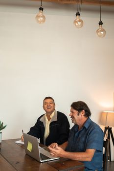 两个成熟的男人在室内对着笔记本电脑大笑，享受着富有成效的团队合作