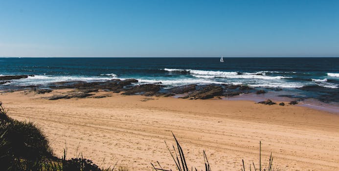 在晴朗的蓝天下，宁静的海滩场景捕捉到了海浪、沙子和远处的帆船
