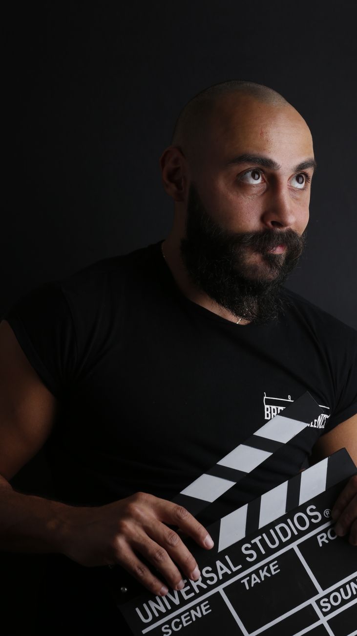 Portret van een kalende man met een baard die een clapperboard vasthoudt tegen een zwarte achtergrond.