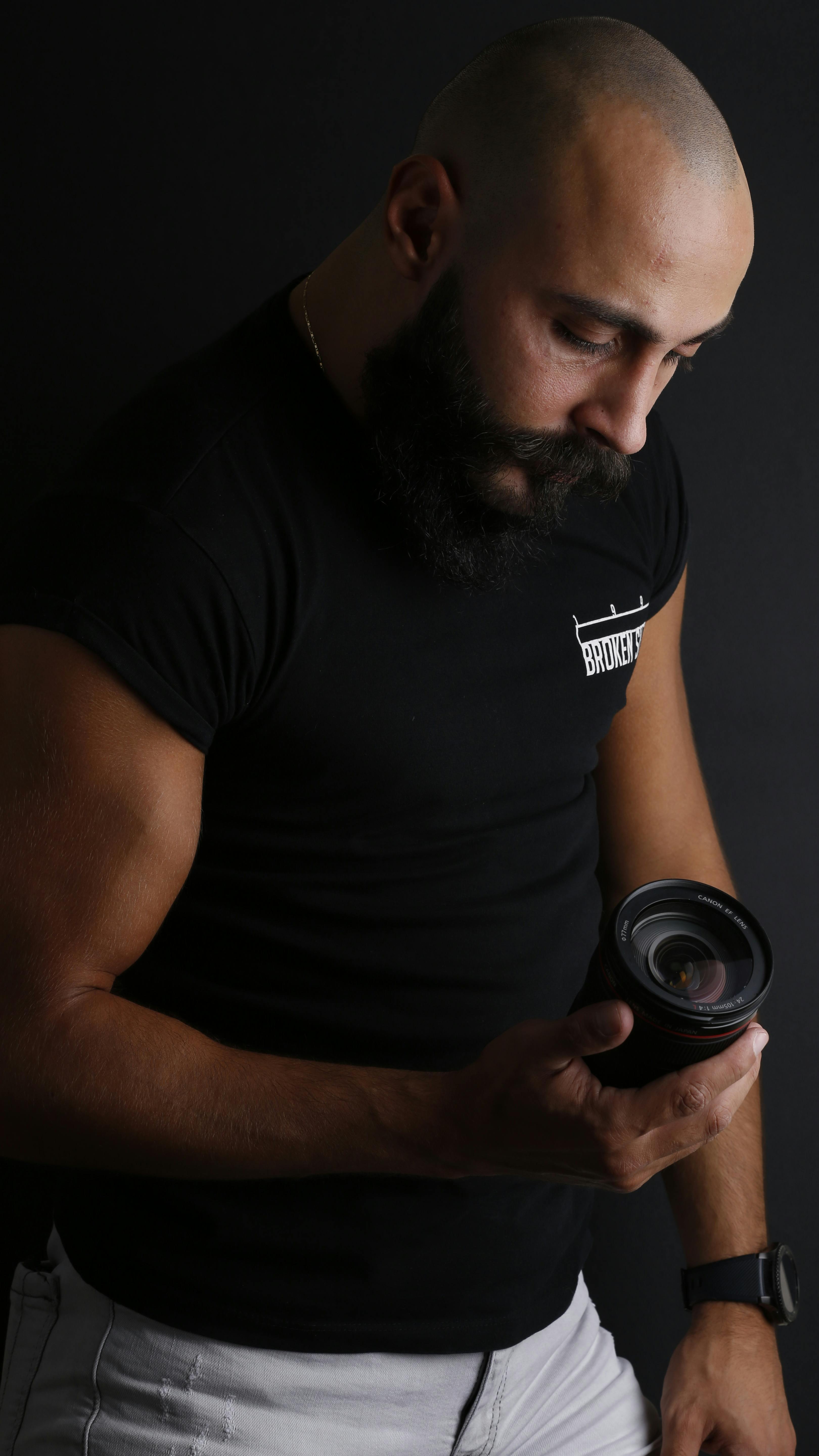 Bald man with beard thoughtfully examining camera lens in a studio setting against a black background.