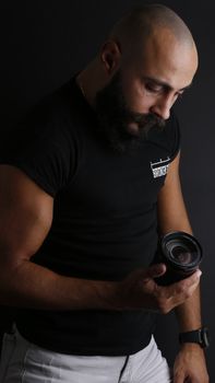 Bald man with beard thoughtfully examining camera lens in a studio setting against a black background.