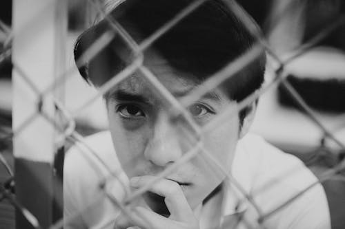 Pensive young Asian man looking at camera through metal net fence