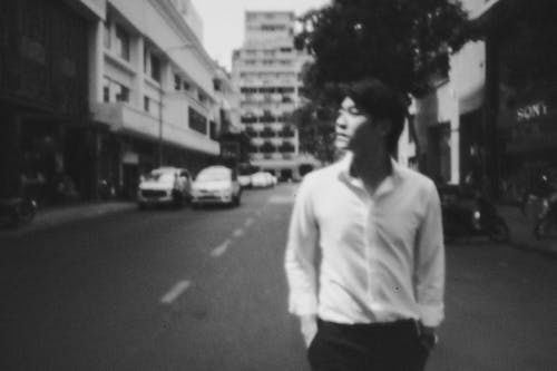 Black and white of young serious Asian man in elegant shirt standing on street near cars with hands in pockets and looking away